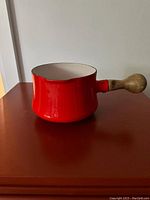 Side view of red butter warmer with wooden handle on dark surface, showing glossy enamel exterior and rounded wooden handle.
