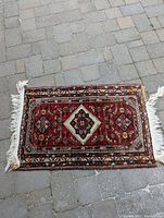 Photo of vintage small rug laid on stone pavement showing vibrant red and multicolor geometric floral design with damaged fringe on one end
