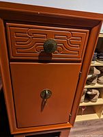 Front view of the cinnamon-colored end table showing the drawer with decorative handle and lower door with a latch, hinged on the right side. Surface shows minor paint chips.