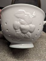 Close-up of the raised berry and leaf pattern embossed on the white ceramic strainer exterior with drainage holes visible.