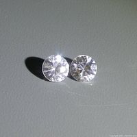 Two round cut colourless natural zircon gemstones displayed on a grey surface showing clarity and cut.