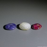 Three oval cabochon star sapphire stones in grey, blue, and pink, shown together on a plain background to illustrate size and color range.