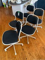 Set of five white metal framed office chairs with black upholstered seats and backs. All chairs shown grouped together on wooden floor.