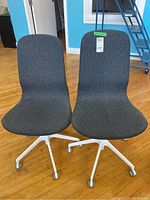 Two dark gray upholstered office chairs with white metal legs and caster wheels on wooden floor, positioned side by side.