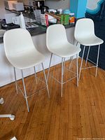 Set of 3 white faux leather bar stools with metal frames placed side by side in kitchen setting on wooden floor.