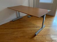 Image showing left side view of wooden desk top with silver adjustable metal frame against wall on hardwood floor.