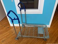 Front angle view of the steel framed dolly showing blue handles and metal mesh platform on caster wheels.