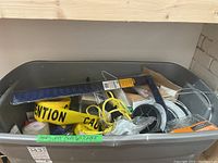 View of gray plastic bin with assorted hardware including yellow caution tape, wire cable, hinges and other items.