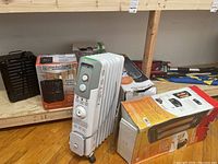 Photo of two Comfort Zone electric utility heaters in boxes and one white fin-type electric radiator heater with control knobs and power switch on wheels.