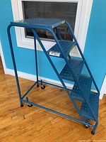 Side view of blue Kleton steel ladder showing perforated steps, rails, and wheels attached at base.