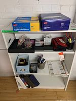 Photo of shelving unit with various office supplies including boxes of envelopes, binders, organizers, pens, and miscellaneous small items.