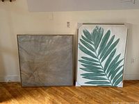 Two framed canvas prints side by side, one with green palm leaf design and one with abstract texture, leaning on a wall on hardwood floor