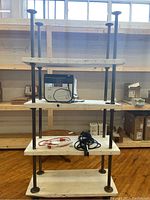 Front view of shelving unit with four white wooden shelves supported by black metal frame on caster wheels. Shows some items on shelves and visible wear.