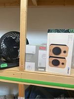 Pair of black Amazon Basics fan, boxed Velux Active unit, and boxed bamboo Bluetooth speakers on shelf