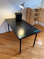 View of black Ikea Linnmon desk with clamping lamp attached and black bin on top, visible scratches and chips on surface.