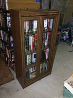 Front and side views of a wooden enclosed bookcase with two glass doors containing books.
