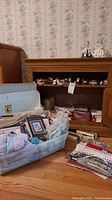 Screenshot of cross stitch supplies in a plastic bin on the floor in front of a wooden cabinet with yarn skeins above it.