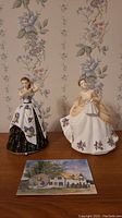 Two Royal Albert porcelain figurines, Linda RA 16 and Margaret RA 13, displayed on a wooden surface against floral wallpaper background. A booklet or pamphlet is laid in front of them likely related to Anne of Green Gables.