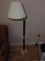 Full view of floor lamp with beige pleated fabric shade, brass tone body, wood checkered pattern on column, placed on wooden floor beside armchair.