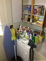 Wide view showing ironing board, duster, tote of cleaning supplies on top of dryer, and open shelves with more items.