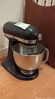 Black KitchenAid Artisan stand mixer with stainless steel bowl on a wooden floor, left side angle view.