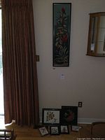 Wide view showing six framed cross-stitch and needlepoint artworks arranged on the floor and hanging on a wall including one large vertical piece with colorful birds.