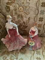 Two porcelain Royal Doulton Pretty Ladies figurines on a patterned couch: Stephanie standing with flowing pink floral dress, Alexandra seated with flower table.