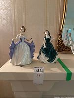 Two porcelain figurines displayed side by side on a mantle against wallpaper background: the larger Scottish Pride on the left and the smaller Irish Charm on the right.