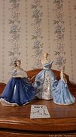 Three Royal Doulton porcelain figurines, Kate, Elaine, and Catherine, arranged on a wooden surface with floral wallpaper background.