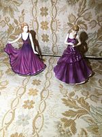 Two Royal Doulton female figurines in purple dresses displayed against a patterned fabric background. The photo shows the front and side views of both pieces with dress details and poses visible.