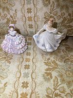 Photo shows two Royal Doulton porcelain ladies figurines side by side on patterned fabric. Kathy in pink layered dress and bonnet, Hannah in white floral gown holding skirt.