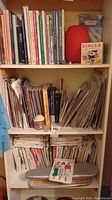 Books and pattern booklets arranged on shelves with a Singer sewing accessory box and red pin cushion.