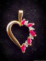 Close-up image of the heart-shaped yellow gold pendant showing the prong-set rubies and diamonds on a black velvet background.