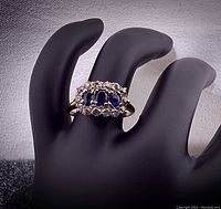 Front view of ring on black hand display showing three oval blue sapphires and surrounding diamonds