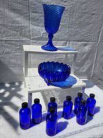 Image showing a blue glass goblet on top shelf, a blue decorative bowl on middle shelf, and nine small blue glass bottles on bottom shelf