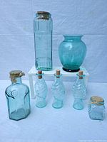 Photo showing seven blue glass containers including a tall rectangular bottle with a glass stopper, a bulbous vase, a rectangular bottle with an embossed circular design, three small ribbed bottles with corks shaped like pineapples, and a small square jar with cork lid.
