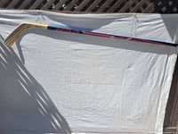 Full length view of a CCM hockey stick laid on a white fabric surface, showing blue and red graphics on the shaft and a natural wooden blade.