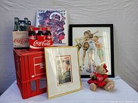 Photo showing vintage Coca-Cola glass bottles in clear and green glass, Coca-Cola bottle crate in red plastic, two framed Coca-Cola prints featuring holiday and sports themes, and a Coca-Cola themed teddy bear.