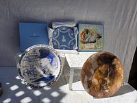 Photo showing all five collectible plates displayed on white chairs, including the Vernon Kilns MacArthur plate, Wedgwood plate box, Disney Peter Pan plate, brown and orange plate, and small blue decorative plate.