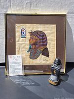 Framed papyrus artwork showing an Egyptian pharaoh's profile with nemes headdress, signed by artist, with small Egyptian-style figurine in front.
