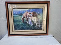 Photo of framed equestrian print depicting a woman standing between a horse and a foal in a meadow with mountains in the background. The frame is wooden with glass front.