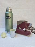 Vintage Stanley film strip projector and thermos with lid and cup shown together on table.