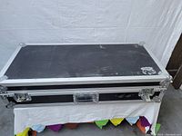 Closed black hard case with metal reinforced corners and edges, latches, and a handle, placed on a white cloth.