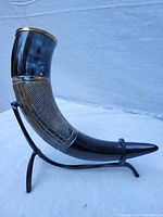 Side view of the drinking horn in the black metal stand showing the polished horn, brass rim, and decorative ribbed metal section with engraved runes.