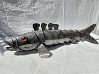 Full view of the metal fish sculpture showing segmented body, fins, tail, and amber eyes against a white textured background.