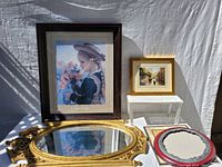 Photo showing framed print of girl with flowers, smaller framed scenic print, oval gold mirror, and empty red and white round frame on white surface with cloth background.