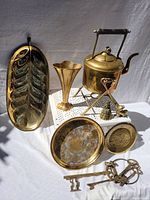 Photo showing a variety of brass items including a leaf-shaped tray, vase, ice bucket with handle, two round trays, small figurines, and two vintage-style keys arranged on a white cloth
