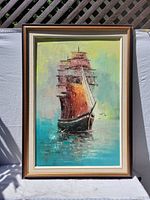 Full framed original oil painting of ship sailing on water with red and white sails and reflections, set against a sky transitioning from yellow to blue, placed outdoors for photo