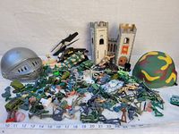 Photo of assorted green and tan plastic army men with helicopters, tanks, two plastic helmets, two castle towers, and various toy accessories arranged alongside a ruler for scale measurement.