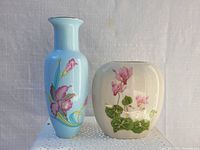 Two porcelain vases showing one tall blue vase with iris design and one rounded cream vase with pink flowers and green leaves, both with glossy finish.